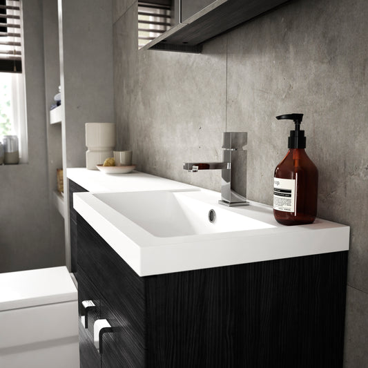 A black vanity unit with white basin and chrome mixer tap, against stone tiles with hand soap on the basin.