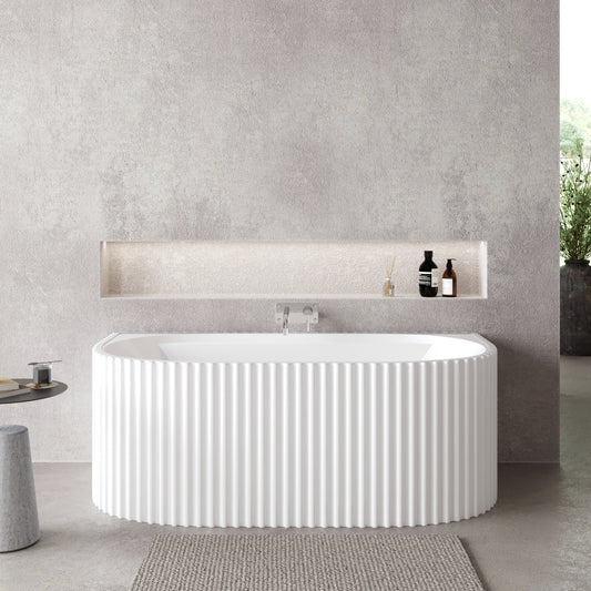 An oval white acrylic bathtub with fluted sides, back wall mounted, with a central waste position, shown in a bathroom setting with minimalistic decor.